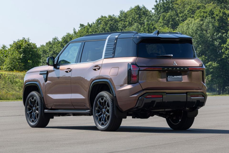 A bronze 2027 Mitsubishi Pajero SUV seen from the rear three-quarter angle on a open tarmac area with trees in the background