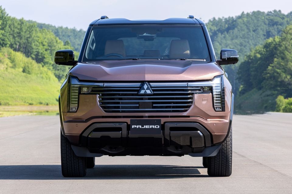 Front view of a brown Mitsubishi Pajero SUV parked on an open road with green trees in the background