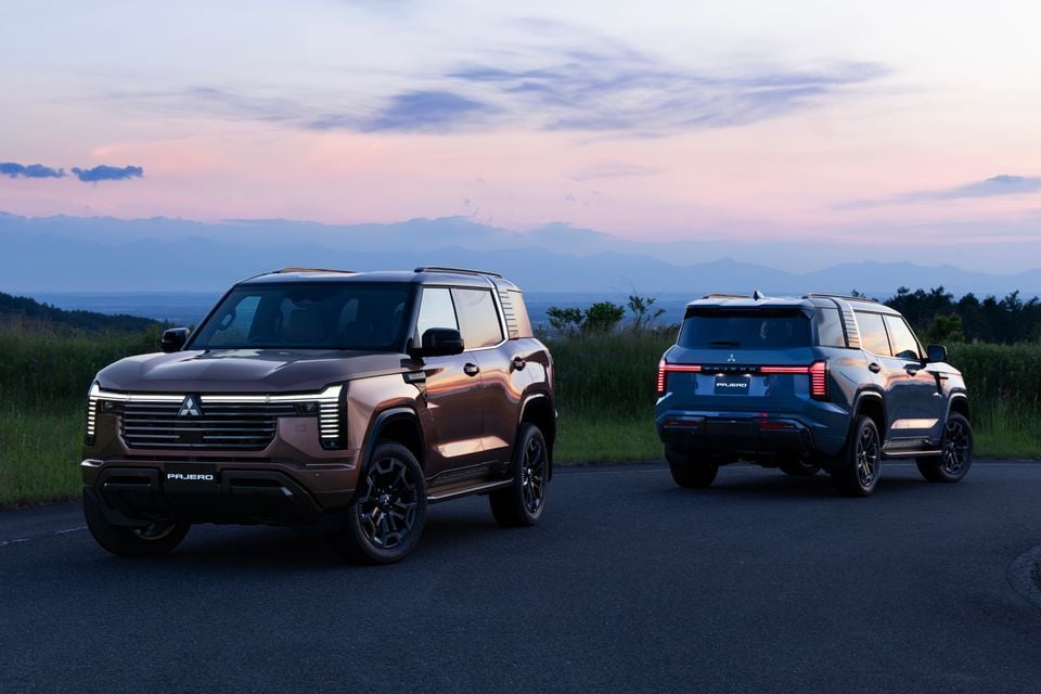 Two Mitsubishi Pajero SUVs parked outdoors at dusk — a bronze model shown from the front and a blue model shown from the rear — with mountains and a pastel sky in the background.