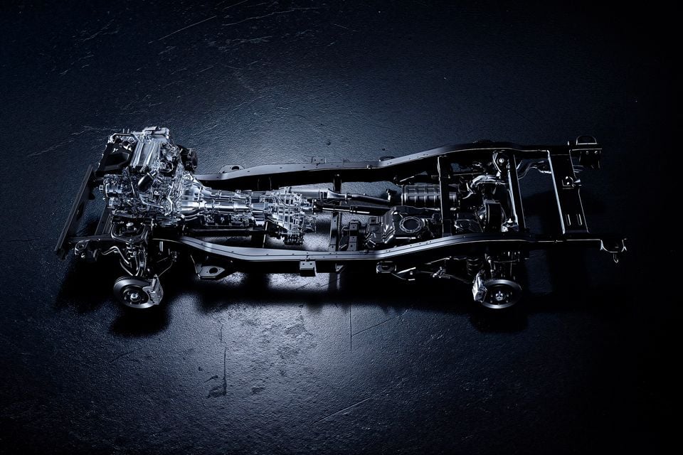 A bare ladder-frame chassis with engine, transmission and drivetrain components displayed on a dark studio surface, seen from above, belonging to the 2027 Mitsubishi Pajero.