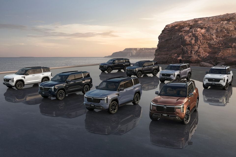 Multiple Mitsubishi Pajero SUVs in various colours posed together on a reflective surface beside a coastal cliff at sunset