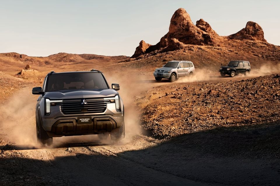 Three generations of Mitsubishi Pajero SUVs driving in convoy along a dusty off-road track through a rocky desert landscape, led by a new-generation Mitsubishi Pajero concept shown from the front.