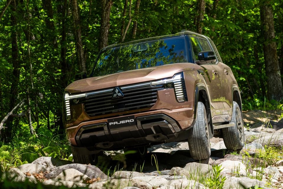 A bronze Mitsubishi Pajero SUV navigating a rocky off-road trail surrounded by forest, seen from the front