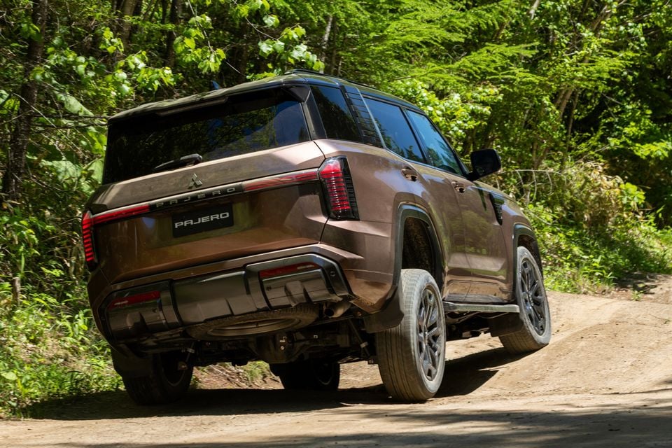 A brown Mitsubishi Pajero SUV seen from the rear three-quarter angle on a dirt track surrounded by trees