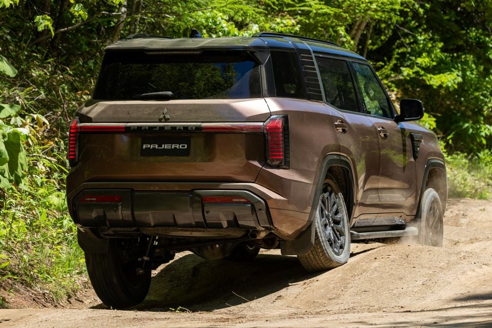 A bronze Mitsubishi Pajero SUV seen from the rear three-quarter angle driving along a dirt trail through a forest