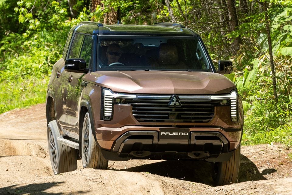A brown Mitsubishi Pajero SUV driving on a dirt trail through a forest, seen from the front