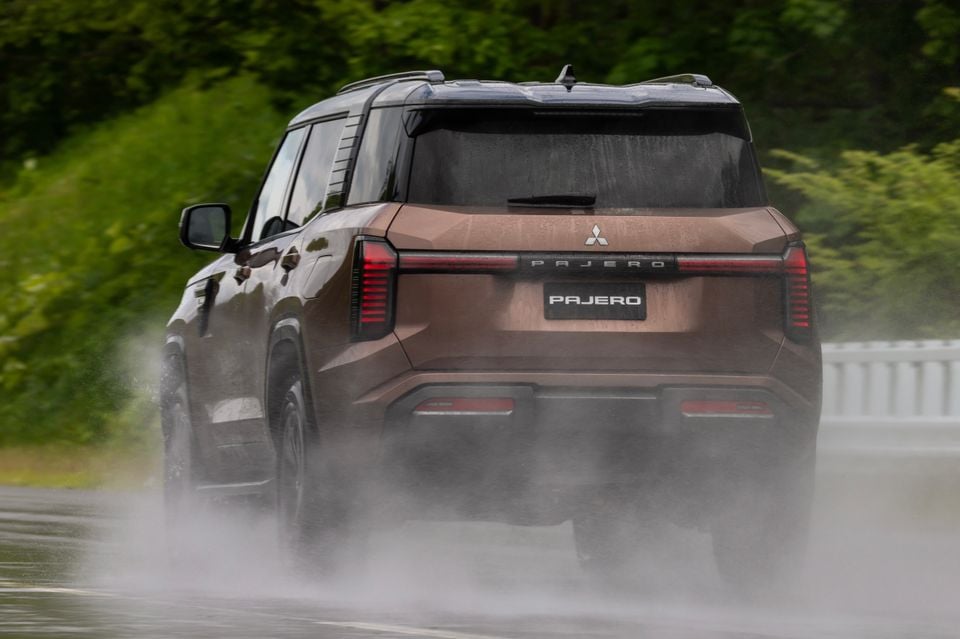 A brown Mitsubishi Pajero SUV seen from the rear, driving on a wet road with spray trailing behind it