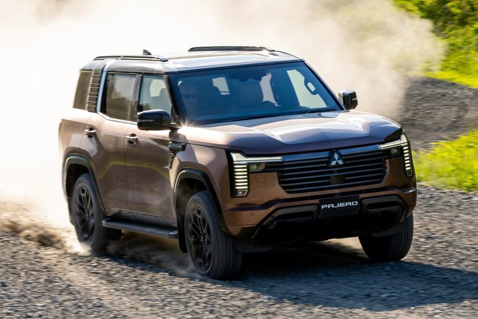A bronze Mitsubishi Pajero SUV driving on a gravel road kicking up dust, seen from the front and side