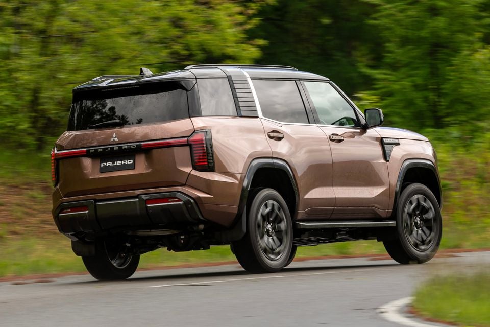 A bronze Mitsubishi Pajero SUV seen from the rear three-quarter angle while driving on a road surrounded by trees