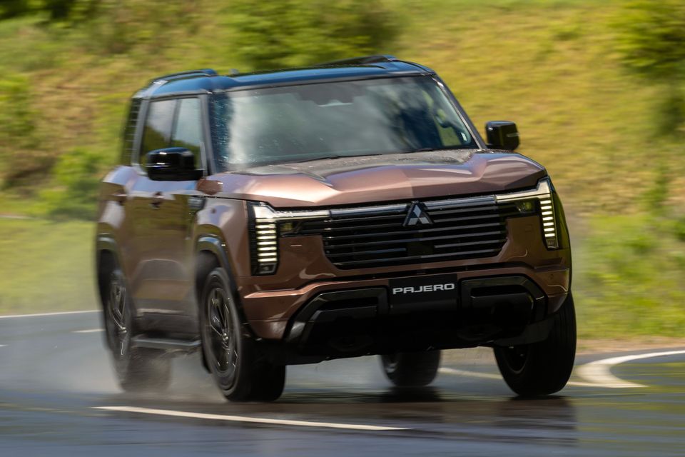 A bronze Mitsubishi Pajero SUV seen from the front driving on a wet road with green vegetation in the background