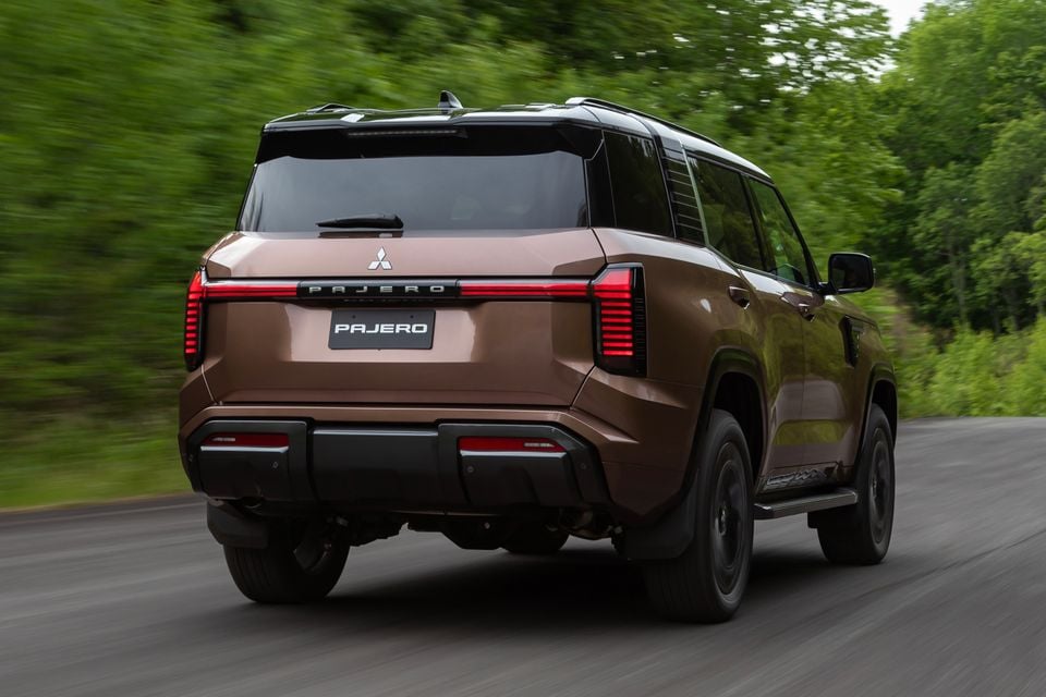 A brown Mitsubishi Pajero large SUV seen from the rear while driving on a road lined with green trees