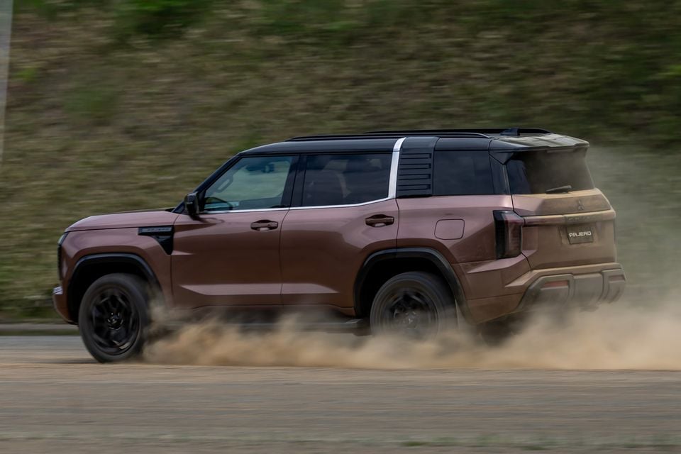 A brown Mitsubishi Pajero SUV seen from the side while driving on a dusty surface, kicking up a cloud of dust