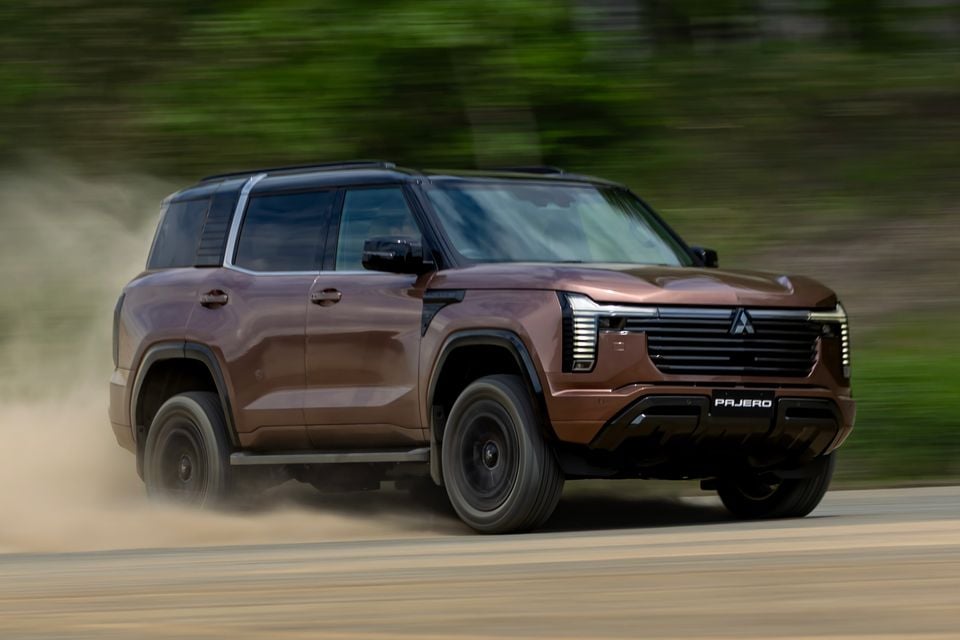 A brown Mitsubishi Pajero SUV driving on a dusty road, seen from the front-side angle with a blurred green background