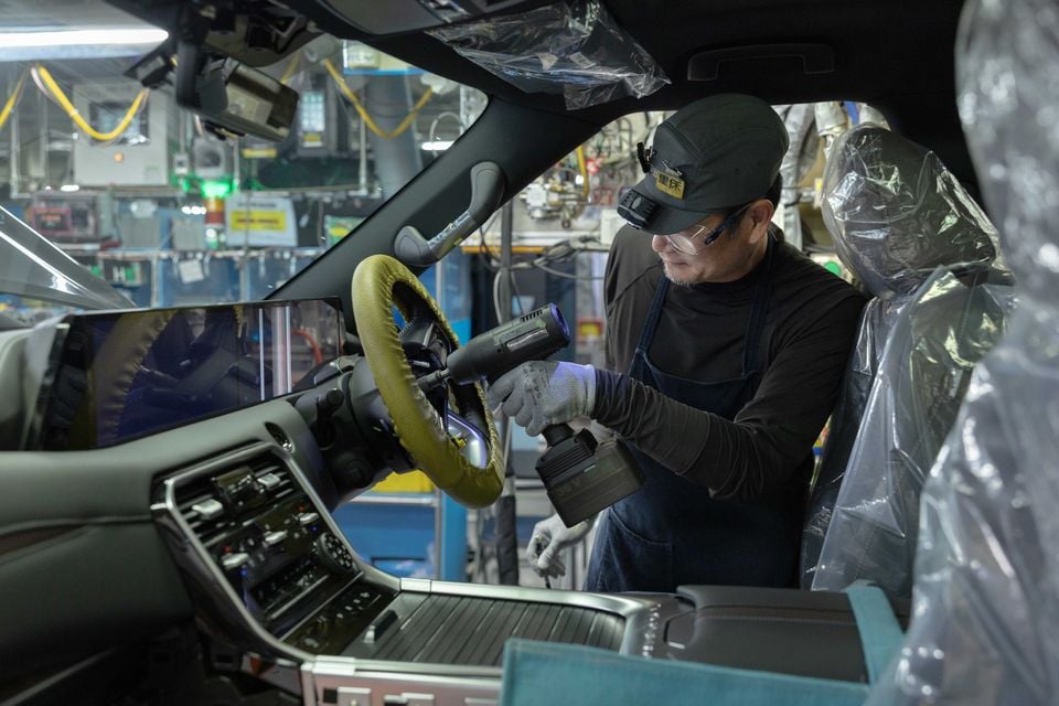 A factory worker uses a power tool to install the steering wheel in a right-hand-drive 2027 Nissan Patrol on the production line