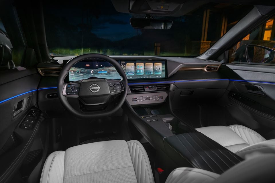 Interior of a 2027 Nissan Rogue Hybrid showing the dashboard, dual widescreen displays, Nissan steering wheel, and light grey leather seats with blue ambient lighting