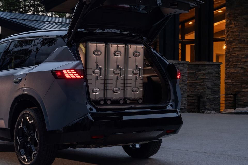 Rear of a grey 2027 Nissan Rogue Hybrid SUV with the tailgate open, showing three large suitcases loaded in the boot, parked outside a modern home at dusk.
