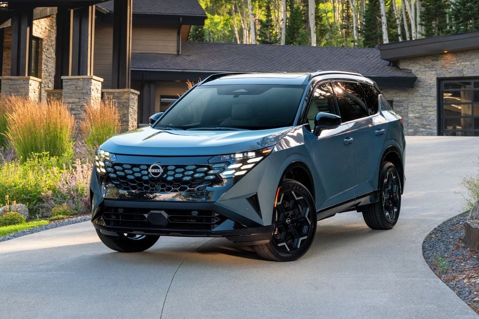 A blue-grey 2027 Nissan Rogue Hybrid SUV seen from the front-side angle, parked on a driveway in front of a stone house.