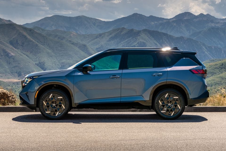 A blue 2027 Nissan Rogue Hybrid SUV seen from the side, parked on a road with a mountain range in the background.