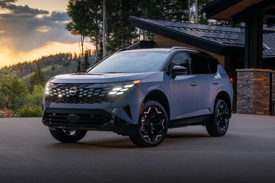 A grey 2027 Nissan Rogue Hybrid SUV photographed from the front-side angle at sunset, parked on a driveway beside a modern stone and timber building.