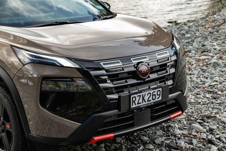 Close-up of the front grille, headlight and Nissan badge on a brown Nissan X-Trail parked on gravel near water.