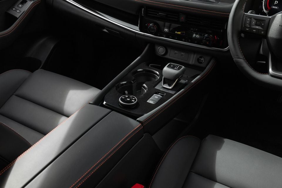 Close-up of the centre console in a 2027 Nissan X-Trail Rock Creek, showing the gear selector, climate controls and cup holders between black leather seats with red stitching.