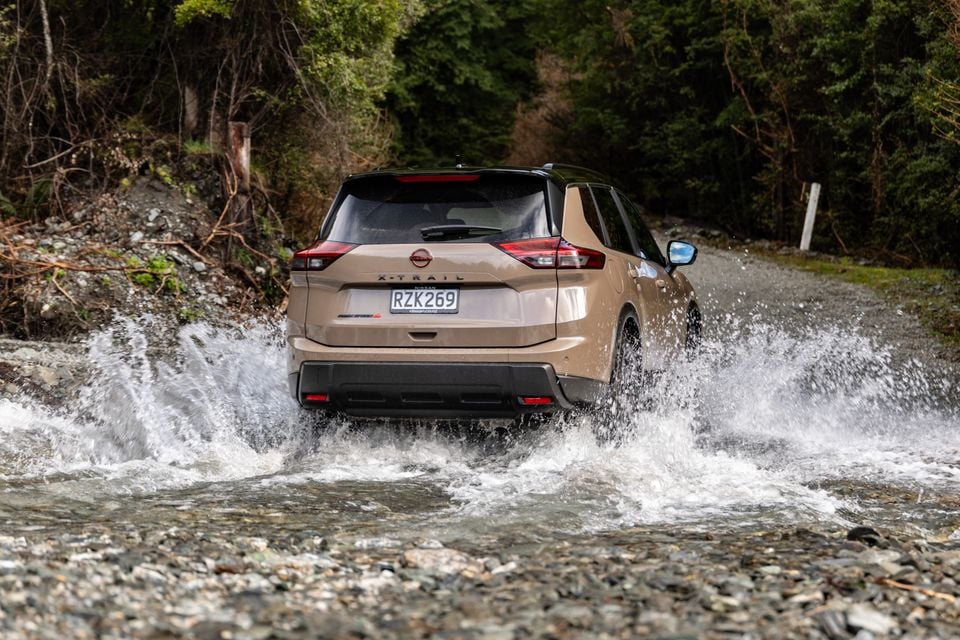 A beige Nissan X-Trail SUV splashes through a shallow creek on a gravel forest road, seen from the rear.