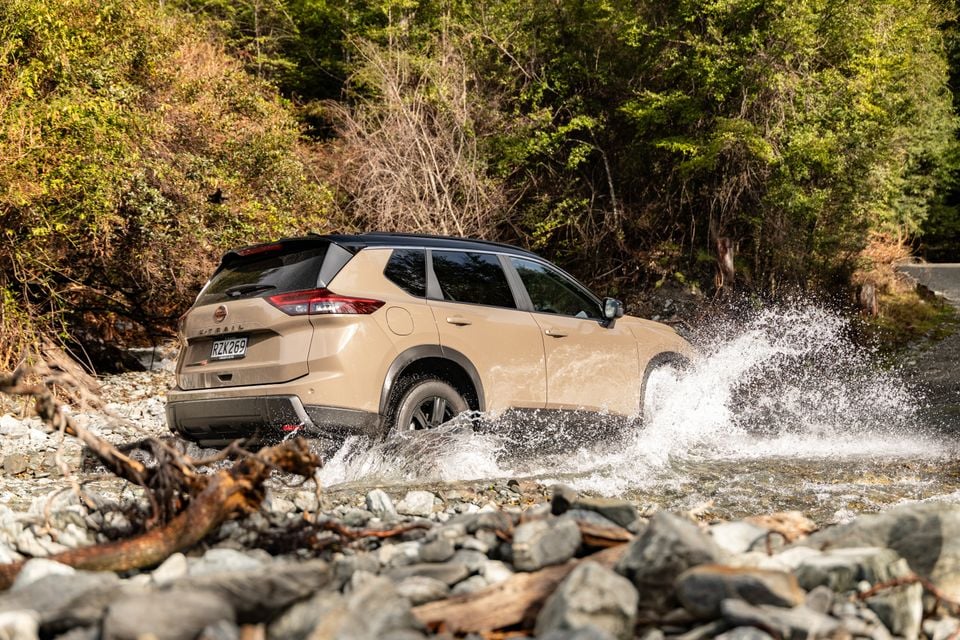 A beige Nissan X-Trail SUV splashes through a rocky creek crossing in a forested area, seen from the rear quarter
