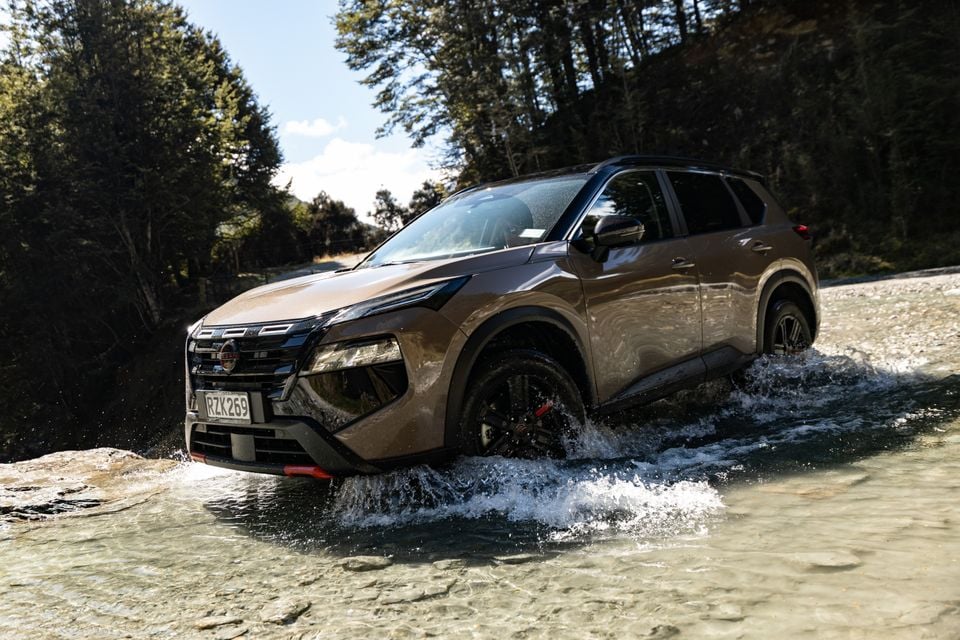 A brown 2027 Nissan X-Trail SUV splashes through a shallow river crossing surrounded by forest.