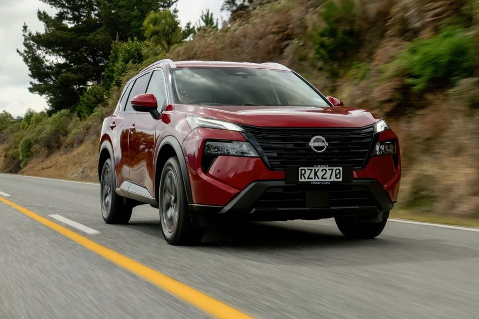 A red Nissan X-Trail SUV driving on a winding road, seen from the front quarter view.