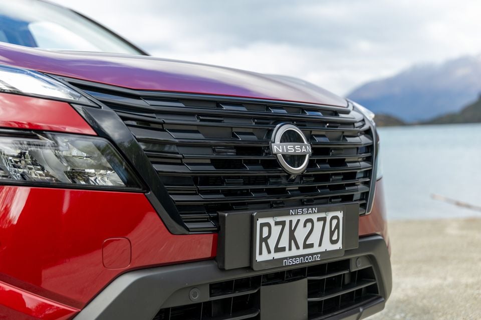 Close-up of the grille and badge on a red Nissan X-Trail, with a lake and mountains in the background.