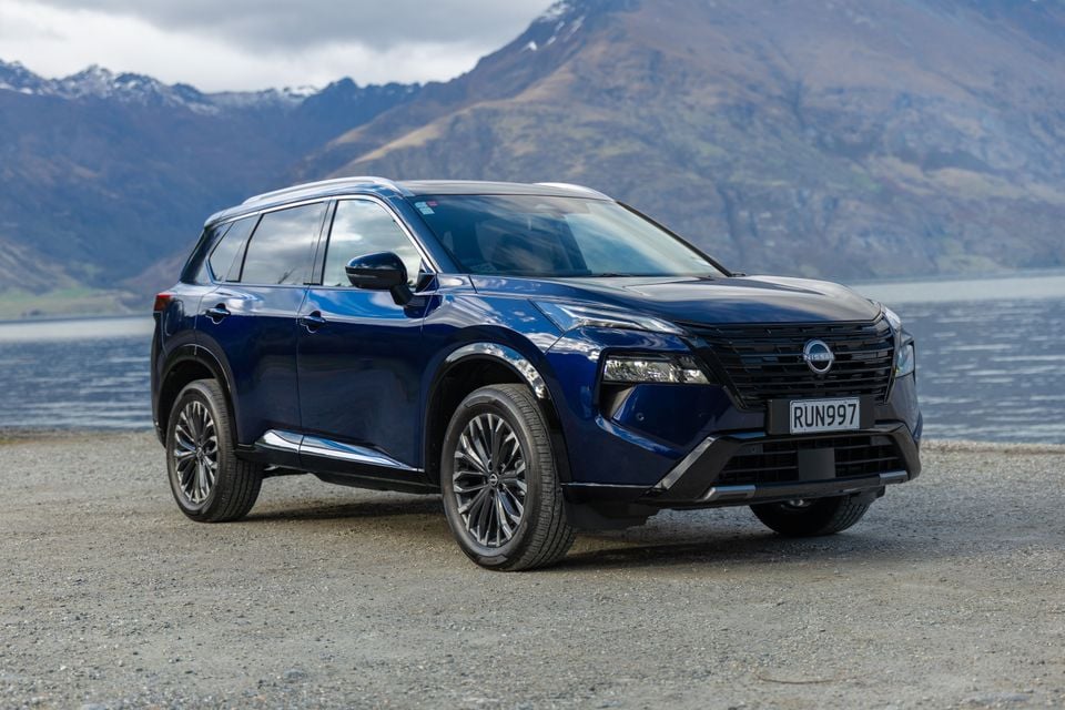 A dark blue 2027 Nissan X-Trail Ti-L e-Power SUV parked on gravel beside a lake with mountains in the background