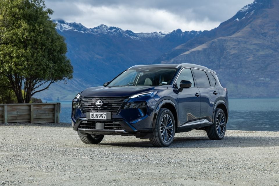 A dark blue 2027 Nissan X-Trail Ti-L e-Power SUV parked near a lake with mountains in the background