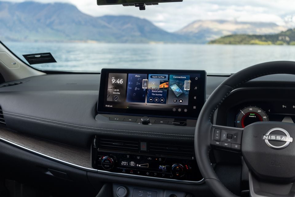 The dashboard of a Nissan X-Trail shows the infotainment touchscreen, climate controls and steering wheel with a lake and mountains visible through the windscreen.