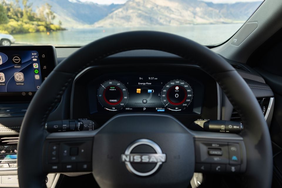 The steering wheel and digital instrument cluster of a Nissan X-Trail e-Power, showing the energy flow display with a lake and mountains visible through the windscreen.