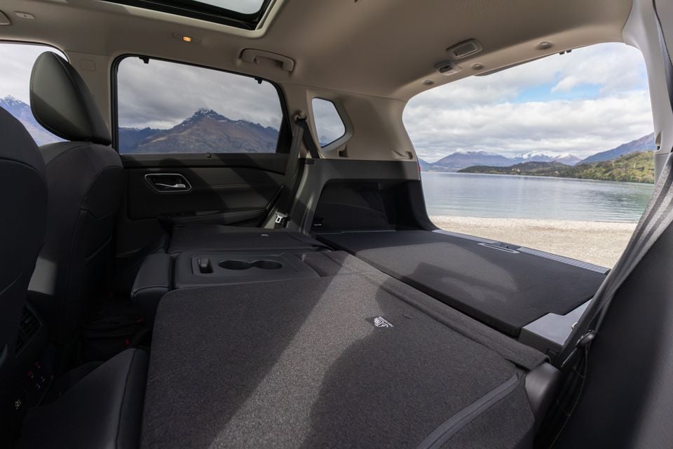 The folded rear cargo area of a Nissan X-Trail SUV, shown with the tailgate open overlooking a lake and mountains