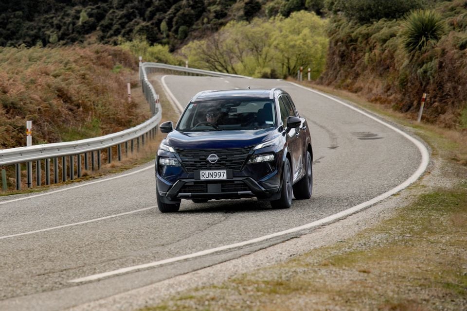A dark blue 2027 Nissan X-Trail Ti-L e-Power SUV driving around a bend on a winding country road.