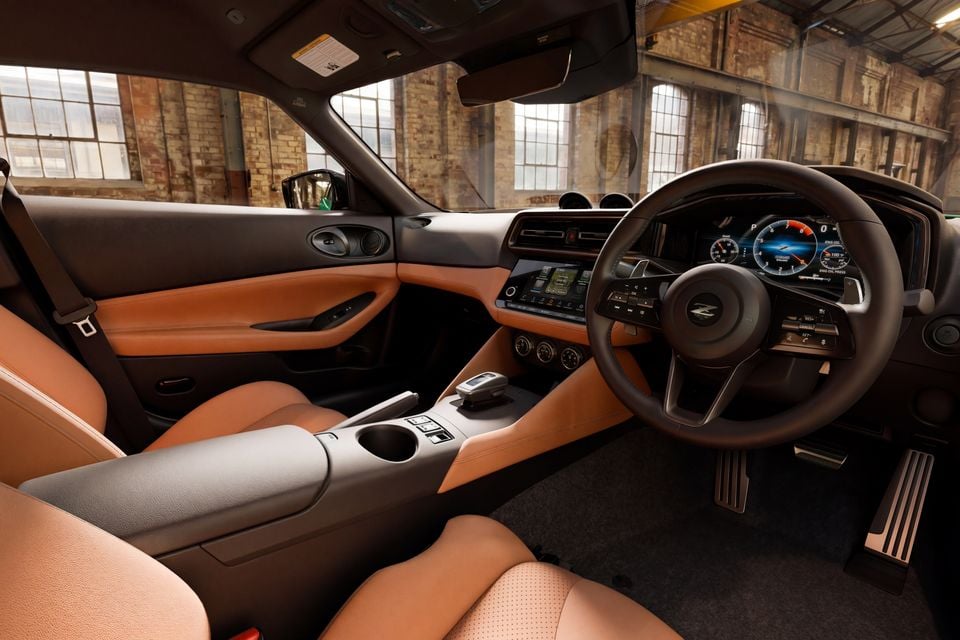 Interior of a Nissan Z showing tan leather seats, dashboard, steering wheel with Z logo, digital instrument cluster, and infotainment screen, photographed inside an industrial warehouse