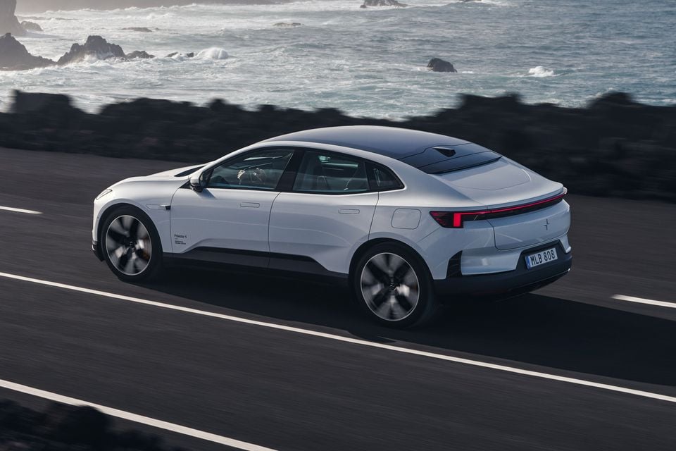 A white Polestar 4 electric SUV coupe driving along a coastal road, seen from the rear three-quarter angle