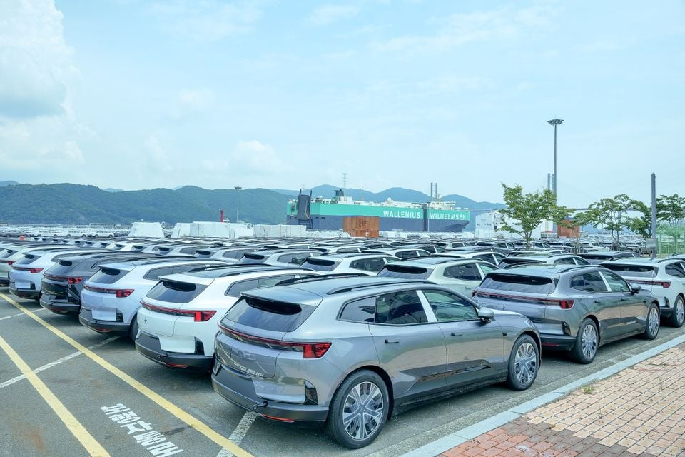 A large group of Polestar 4 SUVs parked at a port awaiting export, with a Wallenius Wilhelmsen car carrier ship visible in the background.