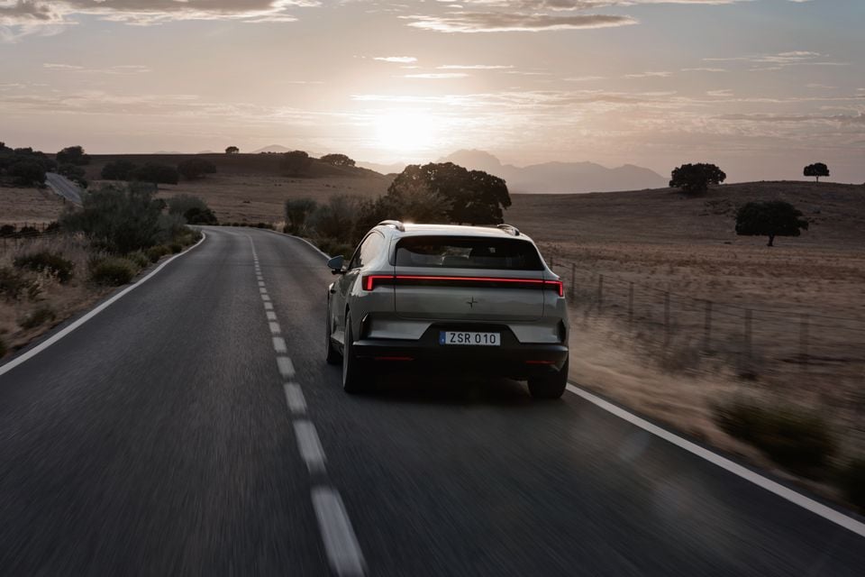A silver 2027 Polestar 4 SUV seen from the rear driving along a rural road at sunset, with hills and trees in the distance