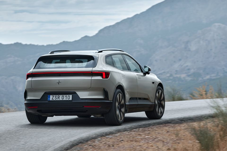 A silver 2027 Polestar 4 SUV driving along a winding mountain road, seen from the rear