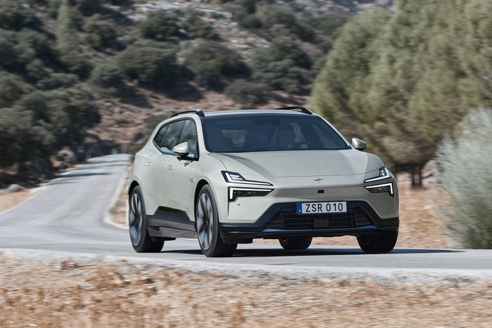 A pale green 2027 Polestar 4 dual-motor electric SUV driving on a winding rural road, seen from the front