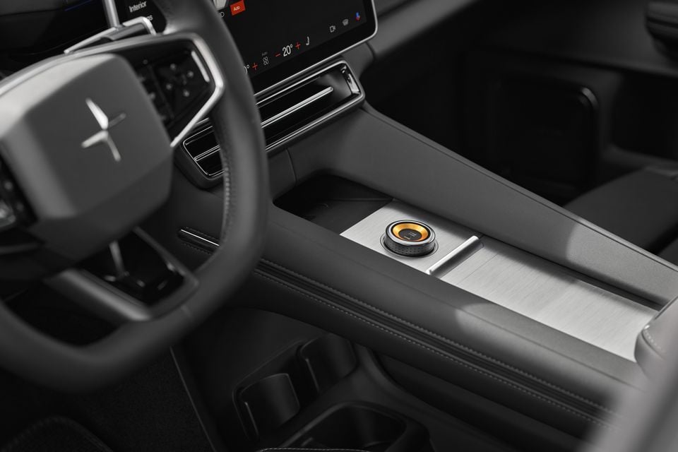 The 2027 Polestar 4 cabin showing the steering wheel, touchscreen and centre console with a brushed metal panel and orange-accented rotary dial