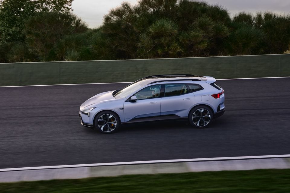 A silver 2027 Polestar 4 electric SUV driving on a race track, seen from the side with a helmeted driver at the wheel