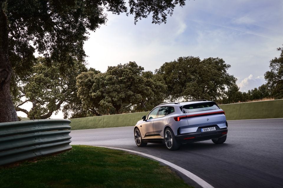 A silver 2027 Polestar 4 electric SUV driving around a tree-lined race track corner, seen from the rear