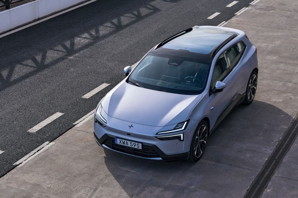 A silver 2027 Polestar 4 SUV seen from above at the front three-quarter angle, showing its glass roof, parked beside a race track