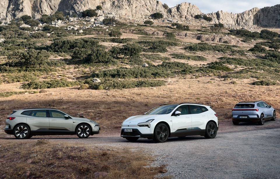 Three Polestar 4 electric SUVs in green-grey, white and silver parked on a gravel road below rocky hills, seen from the side, front and rear