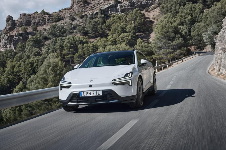 A white 2027 Polestar 4 single-motor electric SUV driving along a winding mountain road lined with pine trees, seen from the front