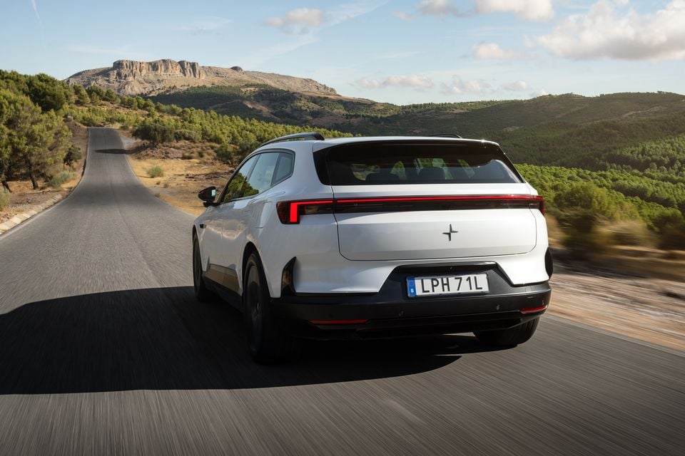 A white 2027 Polestar 4 SUV driving along a winding country road towards rocky hills, seen from the rear
