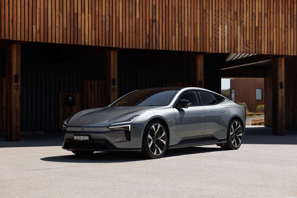 A grey 2027 Polestar 5 Performance sedan parked in front of a timber-clad building, seen from the front left.